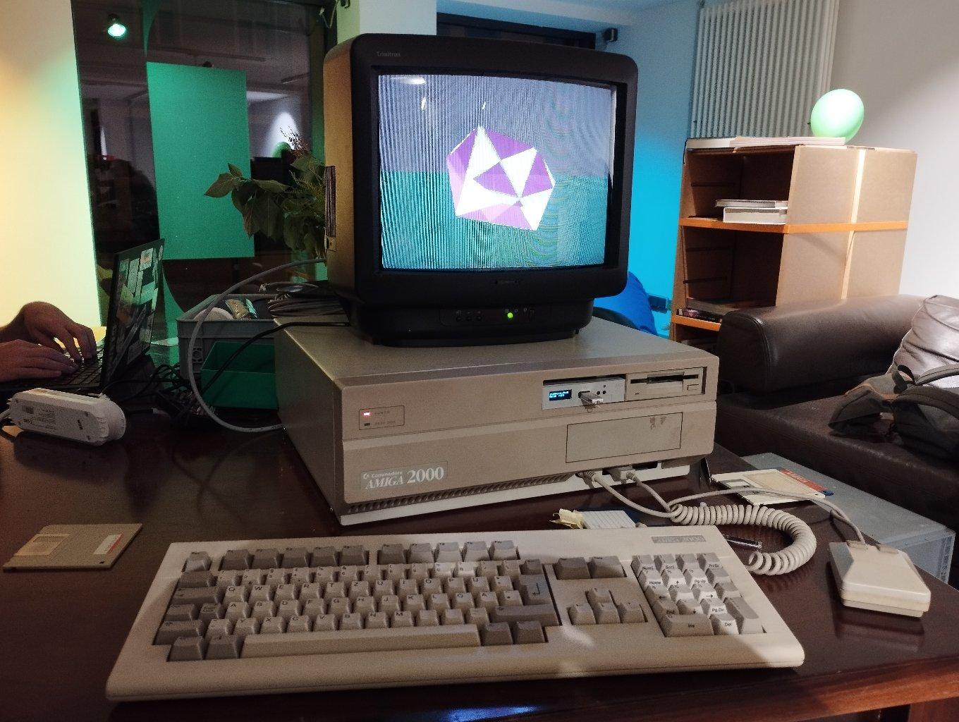 Ein Amiga 2000 mit geöffnetem Gehäuse, und freiem Blick auf die Kabel der Diskettenlaufwerke und das Mainboard. Daneben ein Röhrenfernseher, Tastatur, Maus und Disketten.